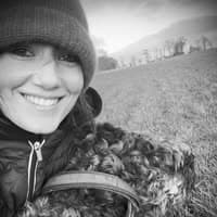 Smiling woman in a beanie and jacket poses with her curly-haired dog outdoors.